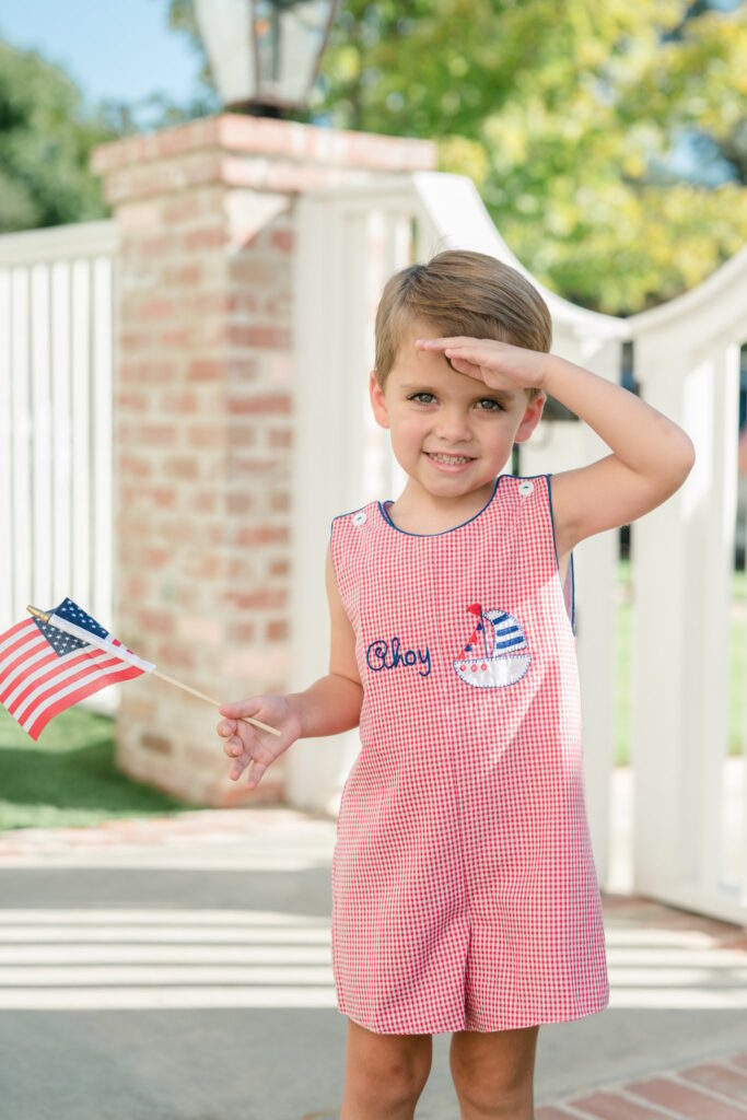 Boy holding a flag wearing a gingham patriotic outfit saluting for a photo.