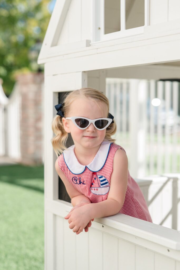 Young girl peering out of a white play hours wearing white sunglasses and a red gingham dress for the fourth of July .