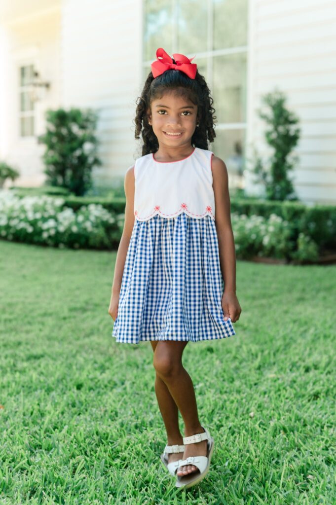 Young girl wearing a blue gingham dress with embroidered fireworks posing for a picture.