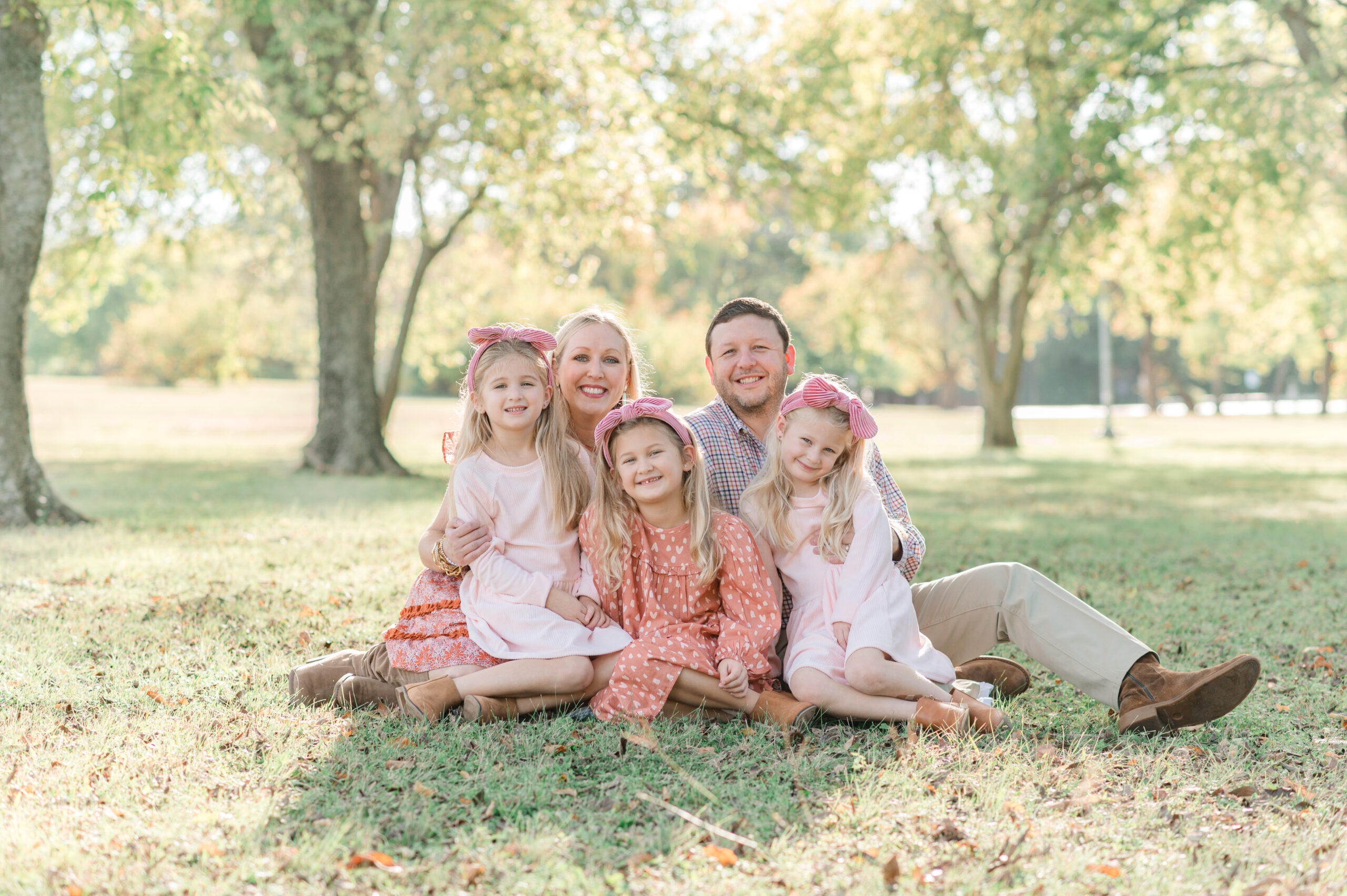 Family portrait by a pro photographer with a mom and dad and three daughters sitting on the grass in a park on a fall day.