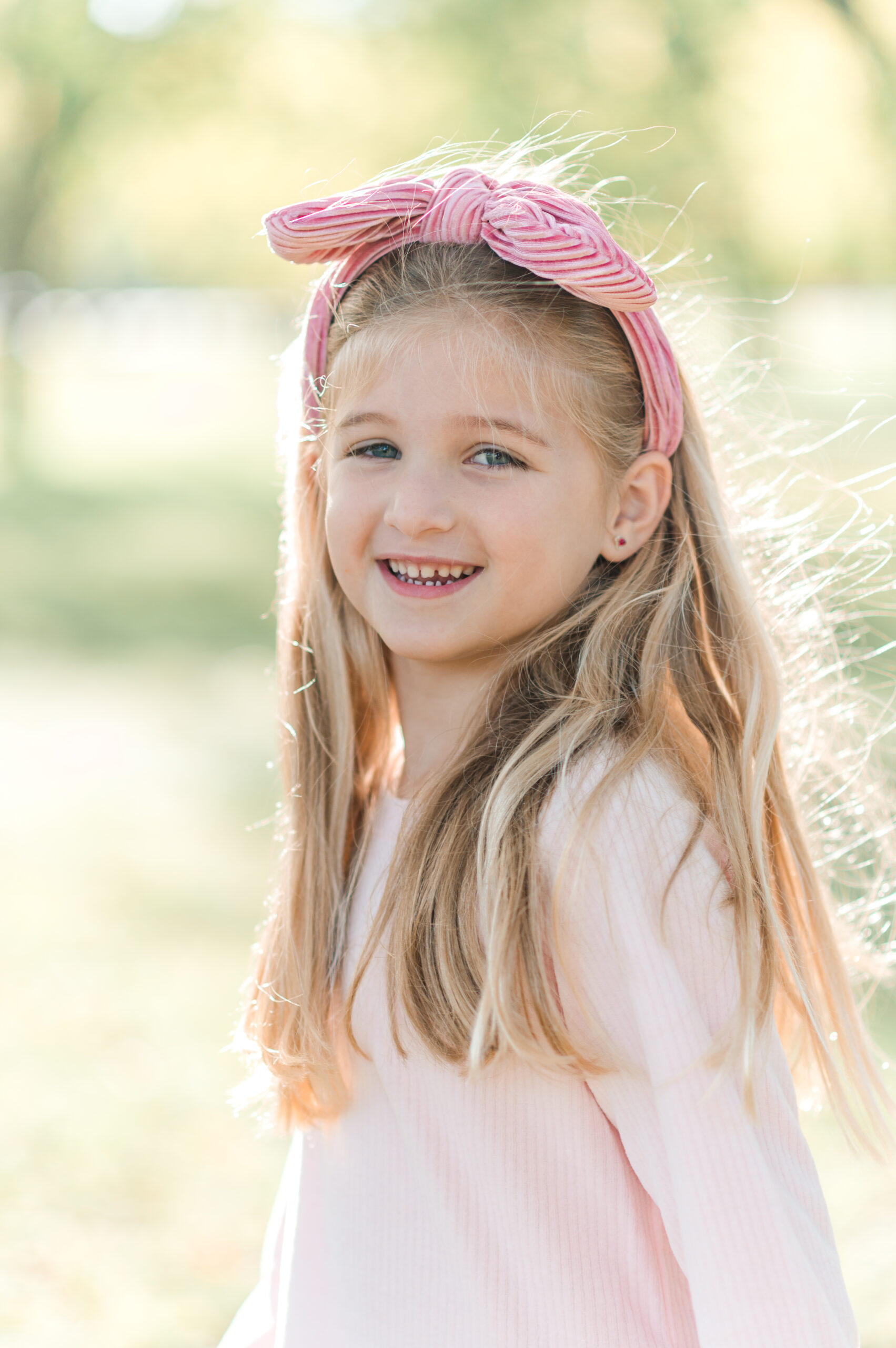 Girl with long blond hair smiling at a pro photographer in a park wearing a pink headband.