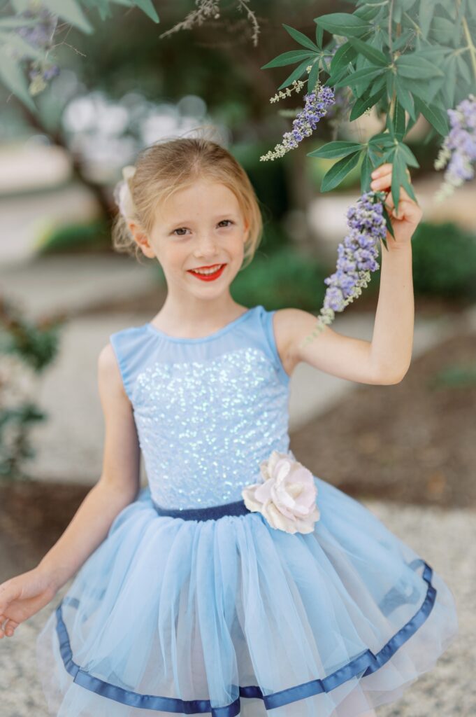 Blond young ballerina wearing a blue tutu and holding a purple flower.