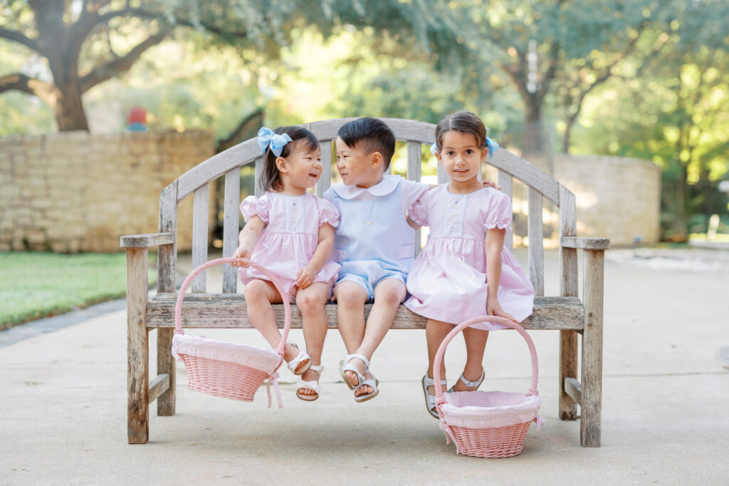 Young boy with his arms wrapped around two of his friends in coordinating pink outfits holding baskets as they prepare for Easter egg hunts in Dallas